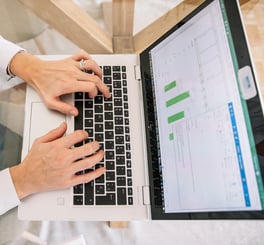  Person typing on a laptop keyboard with an Excel spreadsheet displaying green bar charts